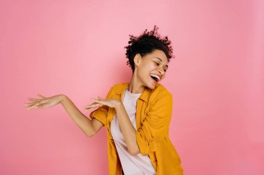Cheerful happy young woman of african american nationality with curly hair, dancing for joy over isolated pink background celebrating success, victory, win, laughing, having fun