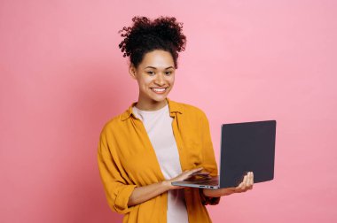 Pretty successful positive african american young curly haired woman, holding an open laptop in hand, looks friendly at the camera while standing against isolated pink background, smiling happily
