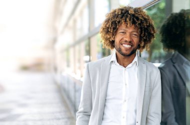 Happy handsome confident positive african american or brazilian man with curly hair, dressed in stylish formal wear, standing outdoors, looking at the camera, smiling friendly. Copy-space
