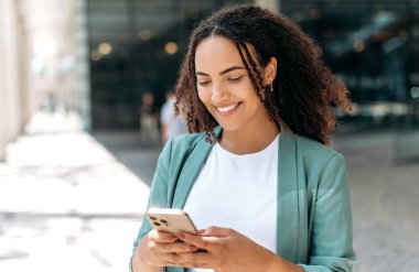 Positive friendly pretty mixed race young woman, with curly hair, stylishly dressed, using her smartphone while standing outdoors, chatting with friends on social networks, answering email, smiling
