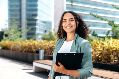 Elegant confident young mixed race woman with curly hair, formally dressed, stands outdoors against the background of the business center, holding a folder with documents, looks at camera, smiles