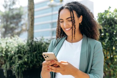 Happy satisfied mixed race young woman, with curly hair, stylishly dressed, using her smartphone while standing outdoors, chatting with friends on social networks, answers email, smiling positively