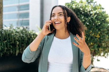 Beautiful positive confident mixed race young woman with curly hair, wearing formal clothes, emotionally talking on a smartphone, standing outdoors, gesturing with her hand, smiling friendly
