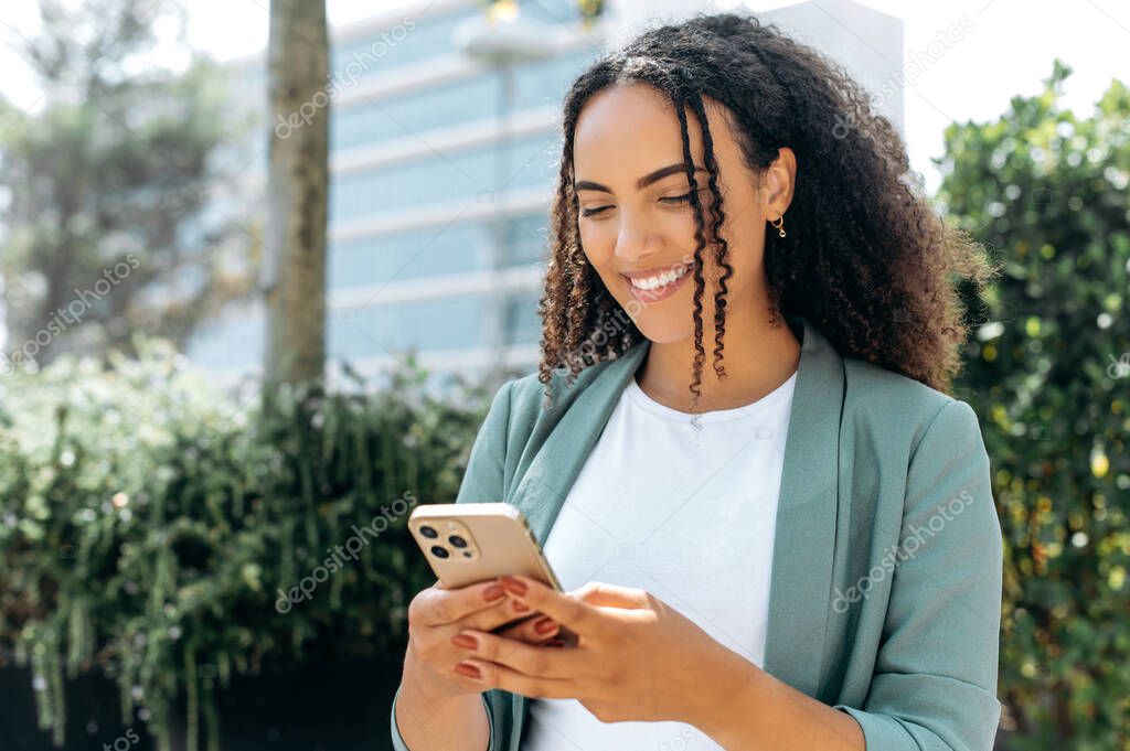 Happy satisfied mixed race young woman, with curly hair, stylishly dressed, using her smartphone while standing outdoors, chatting with friends on social networks, answers email, smiling positively