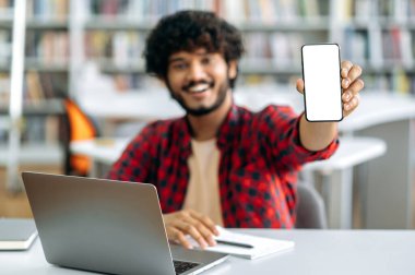 Mock-up and copy-space concept. Photo of defocused Indian or Arabian male student, sitting at table in library, showing his smart phone with blank white screen for advertising and presentation, smile