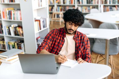 Focused clever arabian or indian college student, sits at table with laptop in university library, preparing for exam, studying information, listening lecture, taking notes in notebook, gain knowledge