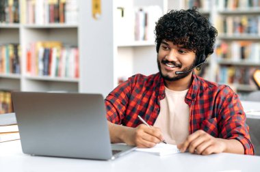 Smart confident positive arabian or indian male student, with headset, sits at table with laptop in university library, preparing for exam, studying information, listens lecture, webinar, taking notes