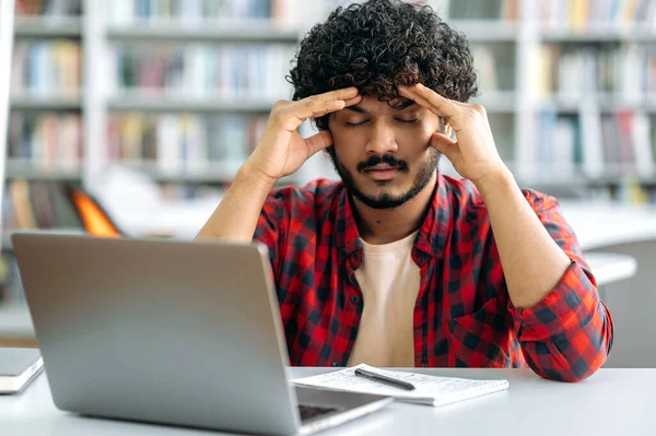 Tired tense indian or arabian guy, university student, wearing stylish shirt, sits at a table in the library, closed his eyes, massages his whiskey, tired of boring studies, has a headache,needs break