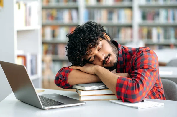 Tired bored arabian or indian university student, is sleeping while sitting at a desk in a library, exhausted of learning, preparing for exam session, suffering of stress and chronic sleep deprivation