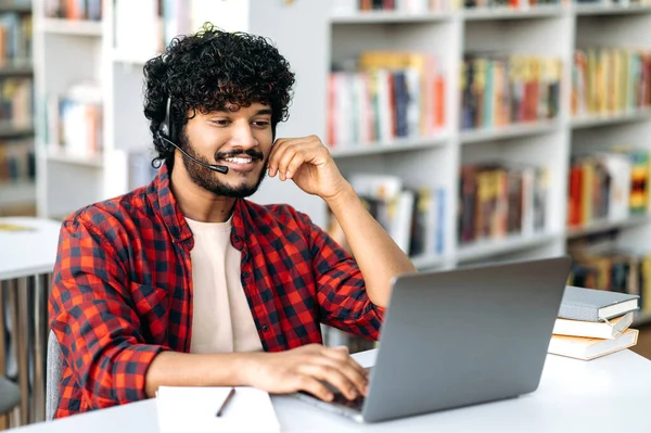 Happy clever university male student, of arabian or indian nationality, with headset, sits at a table with a laptop in the library, studies online, listens to a lecture, webinar, prepares for an exam