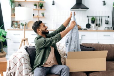Happy smiling indian or arabian guy unpacks his long awaited package, examines the clothes, happy with a good purchase, sits at home on the sofa in the living room. Online shopping concept