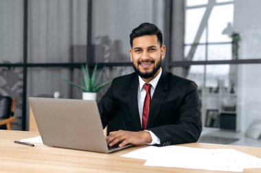 Portrait of handsome confident arabian or indian successful businessman, entrepreneur, lawyer, in a formal suit, sit at work desk with laptop in modern creative office, looks at camera, smile friendly