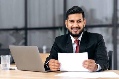 Positive handsome confident arabian or indian successful businessman, lawyer, assistant, in a formal suit, sit at work desk with laptop in modern office, holds document, looks at camera, smiling