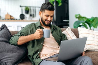 Positive handsome young indian or arabian man in casual clothes, sits on a sofa at home in the living room, using a laptop, browsing the Internet, chatting on social networks, looking at screen, smile