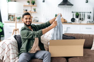 Online shopping concept. Joyful arabian or indian young man unpacking his parcel, happy about getting a long expected order. Mixed race modern guy shopping in internet stores,buying new clothes online
