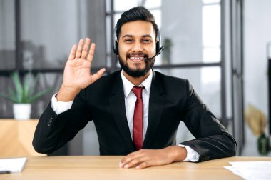 Friendly elegant indian or arabian businessman, financial advisor, sit at desk in modern office, having video call conversation with client, male economic expert records online training course, smiles