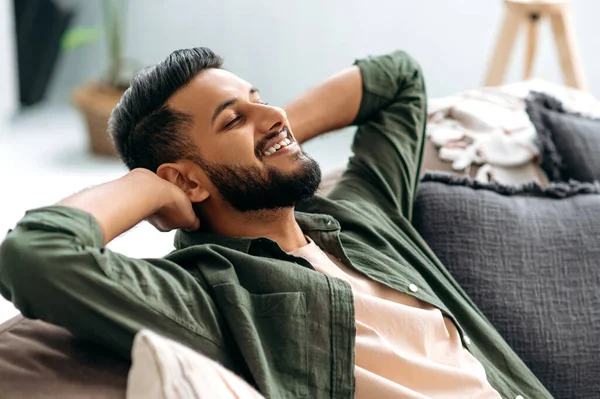 Relaxed satisfied calm mixed race guy in casual shirt, resting on the sofa in cozy living room with his eyes closed, throwing his hands behind his head, dreaming of vacation, resting from work, smiles