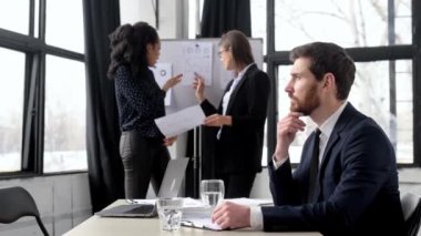 Resmi takım elbiseli başarılı bir iş adamı, iş yerinde oturur, dostça gülümseyerek kameraya bakar ve arka planda iş arkadaşlarının finansal stratejilerini tartışır.