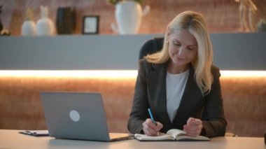 Happy smart caucasian business woman, top manager, ceo, sits at a table in a modern office, uses a laptop, thinks over business strategy and financial opportunities, takes notes, smiles