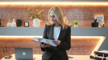 Zeki, kendinden emin orta yaşlı bir iş kadını ya da yönetici ofiste çalışma masasının yanında duruyor, şık bir takım elbise giyiyor, elinde mali belgeler tutuyor, kameraya bakıyor, dostça gülümsüyor.