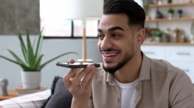 Heyecanlı Hintli ya da Arap bir adam, oturma odasında bir kanepede oturuyor, akıllı telefonunu tutuyor, hoparlörden konuşuyor ve gülümsüyor, sesli mesaj kaydediyor. Kablosuz teknoloji kavramı