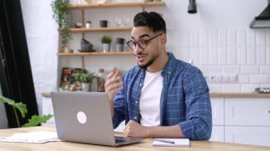Friendly happy arabian or indian man with glasses, sitting at home in the kitchen, conducts video call with client, virtual meeting with colleague, uses laptop, make greeting gesture, smiling