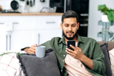 Calm indian or arabian attractive guy using his smartphone while sitting on sofa in living room, browsing internet and social networks, texting with friends or family, holds a cup of coffee in hand