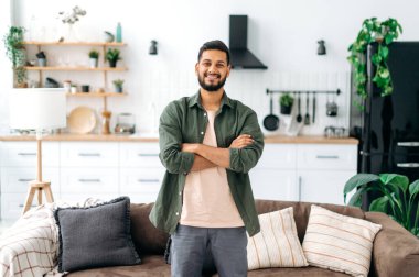 Portrait of a handsome positive modern indian or arabian guy, in casual stylish clothes, standing at home in the living room on the background of the kitchen, with crossed arms, looks at camera, smile