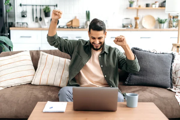 Cheerful amazed arabian or indian guy, freelancer, sits on a sofa in living room, looks at laptop screen, gesticulates with hands, celebrates success, victory, big profit, received good news, smiles