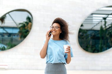 Phone conversation. Busy friendly african american curly haired woman, in formal wear, with glasses, walking outdoors near business center, holding takeaway coffee cup, talking by smartphone, smiling