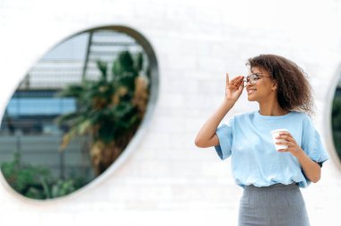 Photo of a positive successful young african american curly woman, wearing blue business shirt, with glasses, standing outdoors near a wall, holding takeaway coffee cup, looks away, smiling friendly