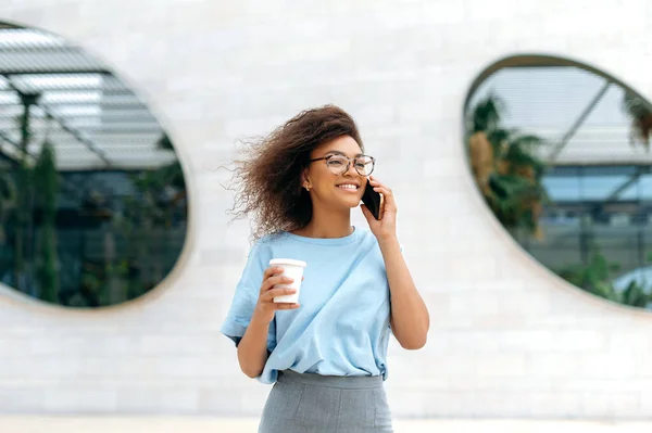 Phone talk, communication. Positive young african american curly woman, in formal stylish wear, with glasses, walking outdoors, holding takeaway coffee cup, talking by smartphone, smiling happily