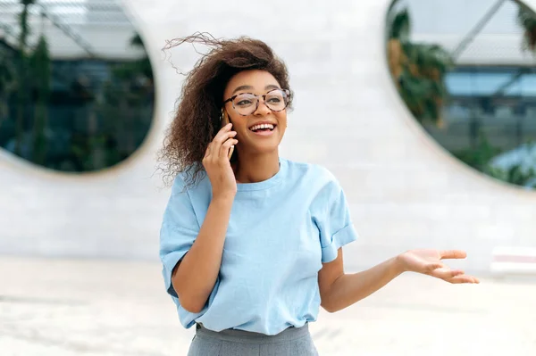 Phone talk concept. Happy young african american curly haired woman, in blue business shirt, with glasses, walking outdoors, going to a meeting, talks by smartphone, looking away, gesture hand, smiles