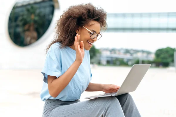 Virtual meeting concept. Busy successful african american young woman, business lady, sitting outdoors, holds online video conference by laptop, discuss financial strategy with colleagues, smiling