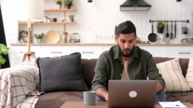 Cheerful amazed arabian or indian guy, freelancer, sits on a sofa in living room, looks at laptop screen, gesticulates with hands, celebrates success, victory, big profit, received good news, smiles