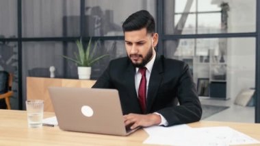 Positive successful young male confident arabian or indian businessman, successful entrepreneur in a suit, sit at a work desk with laptop in modern creative office, looks at camera, smile friendly