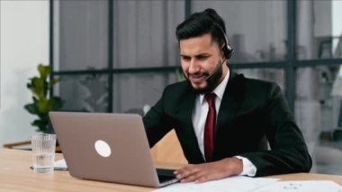Positive successful indian or arabian man in suit, with headset, business advisor, call center worker, company representative, support service operator, holds online consultation, talking with client