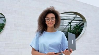 Güzel Afrikalı Amerikalı genç bir kadının portresi. Kıvırcık saçlı, mavi gömlekli, gözlüklü, serbest çalışan ya da öğrenci, dışarıda duran, dizüstü bilgisayarı olan, kameraya bakan, gülümseyen bir kadın.