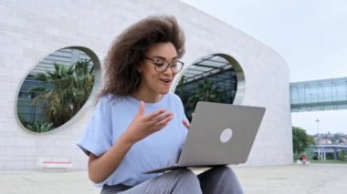 Virtual meeting concept. Busy successful african american young woman, business lady, sitting outdoors, holds online video conference by laptop, discuss financial strategy with colleagues, smiling