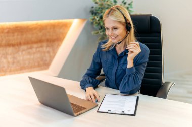 Successful elegant intelligent mature caucasian businesswoman, female financial advisor, in formal wear, with headset, sits in modern office, working in a laptop, holds online consultation, smiling