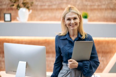 Portrait of an intelligent beautiful mature Caucasian woman, business lady, in business clothes, holding a laptop in her hand, standing in the office near the desktop, looking at the camera, smiling