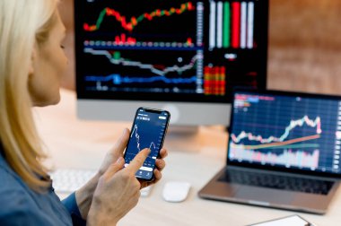 Smart phone with stock diagrams on screen in female hands.Female crypto trader investor broker using cellphone and laptop for cryptocurrency financial market analysis, buying or selling cryptocurrency