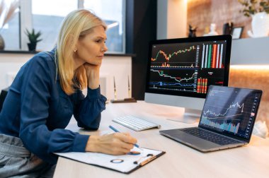 Focused clever mature caucasian smart blonde woman, a stock trader, a crypto investor, sit a the desktop, analyzes charts on a computer and laptop screen, compares indicators, assesses risks