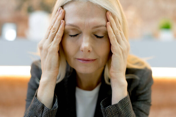 Крупным планом уставшая белая деловая леди средних лет, испытывающая усталость от работы, переутомление, головную боль, мигрень, массирует виски, закрывает глаза, нуждается в отдыхе