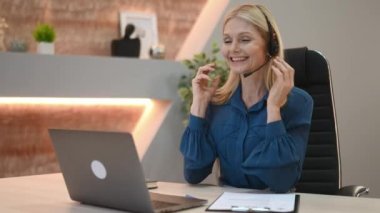 Positive elegant mature caucasian businesswoman, female call center operator, financial advisor, in formal wear, with headset, sits in modern office, holds online consultation with client, waves hand