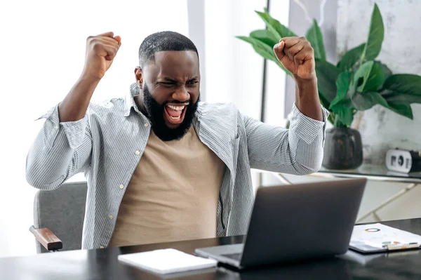 Amazed happy emotional african american stylish man, trader, student, freelancer, sitting at table with laptop in the office, enjoying big profit, victory, good news, gesturing with fists, smiling