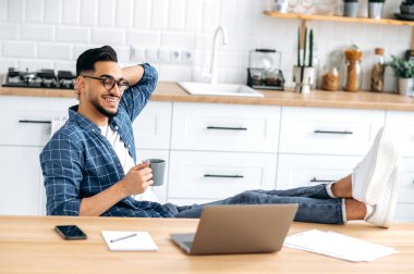 Relaxed indian or arabian modern guy in stylish clothes, sit at home in a kitchen with his legs on the table, holding a cup of coffee in hand, working remotely from home using laptop, chilling, smiles