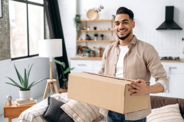 Happy confident indian or arabian guy, stand at home in living room, holding a large cardboard box, received a long-awaited parcel from the online store, preparing to unpack, looks at camera, smiles