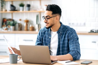 Job with documents. Smart successful arabian or indian guy with glasses, freelancer or office worker working remotely, sit at a desk at in s kitchen, working on a project, analyzing documents, smiling