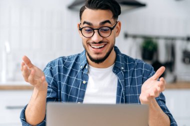 Excited positive Arabian or Indian man with glasses, clever student or freelancer, having distant communication by video call uses laptop, listening to an online webinar, lecture, smiling friendly